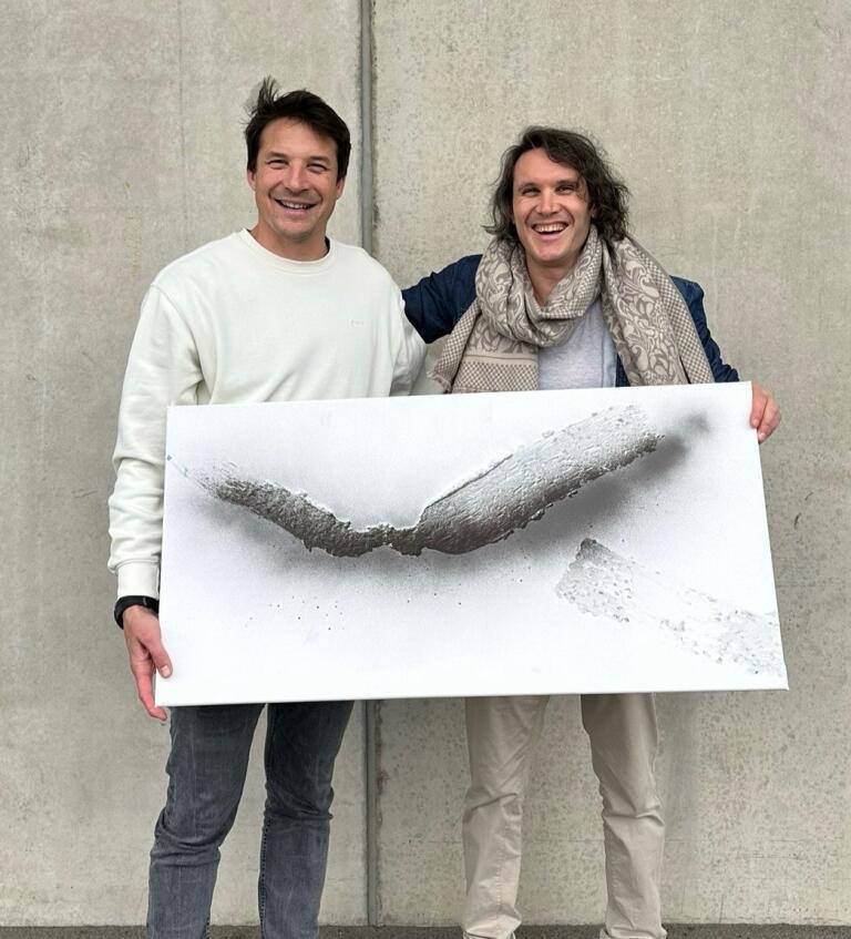 Werner Wilfred tenant une toile en relief blanc avec un collaborateur — échelle et matière de l'œuvre.